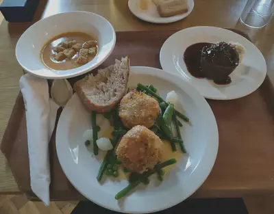 A white plate with a slice of bread and two round fishcakes on a bed of green beans, peas, and pickled white onions. Also a bowl of soup with croutons and a small white plate holding a small pudding with sauce.