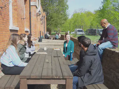 6 people at a picnic table talking. Most are looking to a man sitting on a low brick wall behind one of the table benches. More people talking at a similar table in the background.