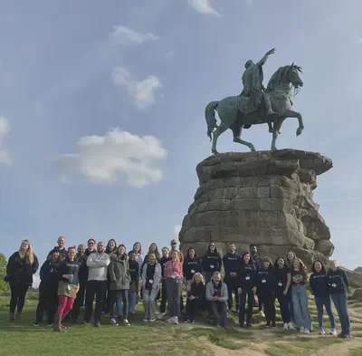 Group of 29 people in front of a copper statue of a man on a horse. The statue has a green tinge and is on top of a pile of stone.