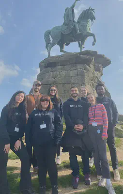 Group of 8 people in front of a copper statue of a man on a horse. The statue has a green tinge and is on top of a pile of stone.