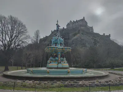 A blue fountain with gold and brown details, topped with a woman holding a sceptre. Edinburgh Castle stands atop Castle Rock in the background.