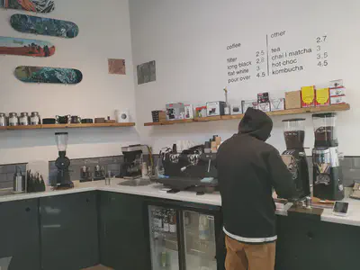 The interior of a coffee shop with skateboard decks on the left wall and the drink menu on the right wall. The foreground shows shelves with coffee equipment above a counter top that boasts 3 coffee grinders, a 2-group espresso machine, a batch brewer, a sink, and various other coffee things. A fridge is built into the storage space below the counter.