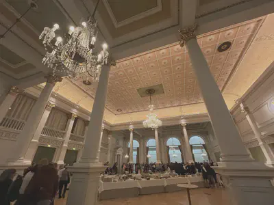 Large elegant room with perspective slightly distorted due to zooming. Glass chandeliers hang from a patterned ceiling. There are large white columns in the foreground and many people line the walls of the room. Two buffet tables full of food are in the centre of the room.