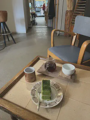 A rectangular slice of green two-layered cake on a rose decorated plate, a bronze coloured cup of water, and a carafe of coffee with a white cup on a wooden board, all atop a brown and beige table. Across the table is a wooden chair with blue cushions.