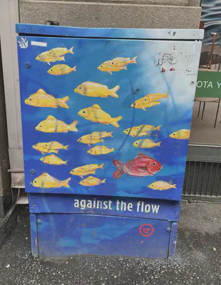 An electrical box on the street painted with a blue background and numerous yellow fish facing left. One red fish is facing right. At the bottom of the box are the words against the flow.