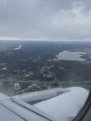 Overhead view of Helsinki from an airplane window with part of the aircraft visible at the bottom of the photo. Top of the photo is full of clouds with the middle showing highways, large water bodies, and a lot of thick greenery. Buildings appear as small shapes.