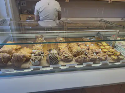 Glass display of assorted viennoiserie. From left to right, large plain croissants, croissants topped with cheese wrapped in parchment paper muffin cases, pain au chocolat, cinnamon buns, croissants with cookie topping, square shaped kouign amann on parchment paper, and pasteis de nata. A person in a white tshirt stands behind the display case with their back facing the camera.
