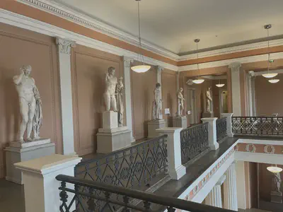 Five sculpted statues line the left wall, which is painted a soft brown and portioned by grey columns. Each portion has a textured border and each statue is in the centre of a portion. The walkway in front of the wall serves as one border of an open space in the floor which is blocked by ornate black railings. Hemispherical lamps hang from the ceiling.