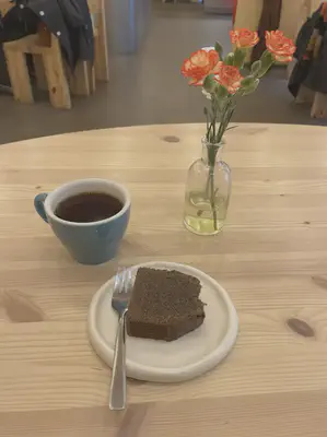 A table with horizontal light stained wood grain. In the foreground is a slice of brown loaf cake with a small metal fork on a white ceramic plate. On the top left of the plate is a blue cup of black coffee and to the top right is a transparent glass bottle serving as a vase for orange and white flowers.