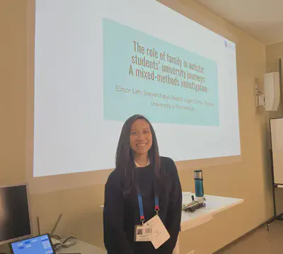 An Asian woman with long brown hair and wearing a navy blue sweater with conference lanyard around her neck stands in front of two desks which are against a wall. The title slide of a presentation is projected onto the wall. Slide title is The role of family in autistic students&rsquo; university journeys colon a mixed methods investigation. Slide subtitle is author information which reads Elinor Lim, Steven Kapp, Beatriz Lopez, Emine Gurbuz, University of Portsmouth.