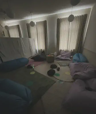 Dark unlit room with blue and purple beanbags and round black cushions on the floor. The floor is also littered with rugs and stim toys. Small glow of light from a green fibre optic toy in the centre left of the photo and a wall of white cloths behind it.