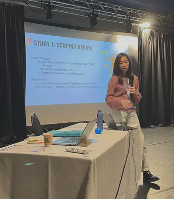 An Asian woman with long, dark brown hair wearing a pink sweater, light blue jeans, and black boots, standing in front of a projector screen and holding a microphone. She looks at the laptop on the table in front of her.